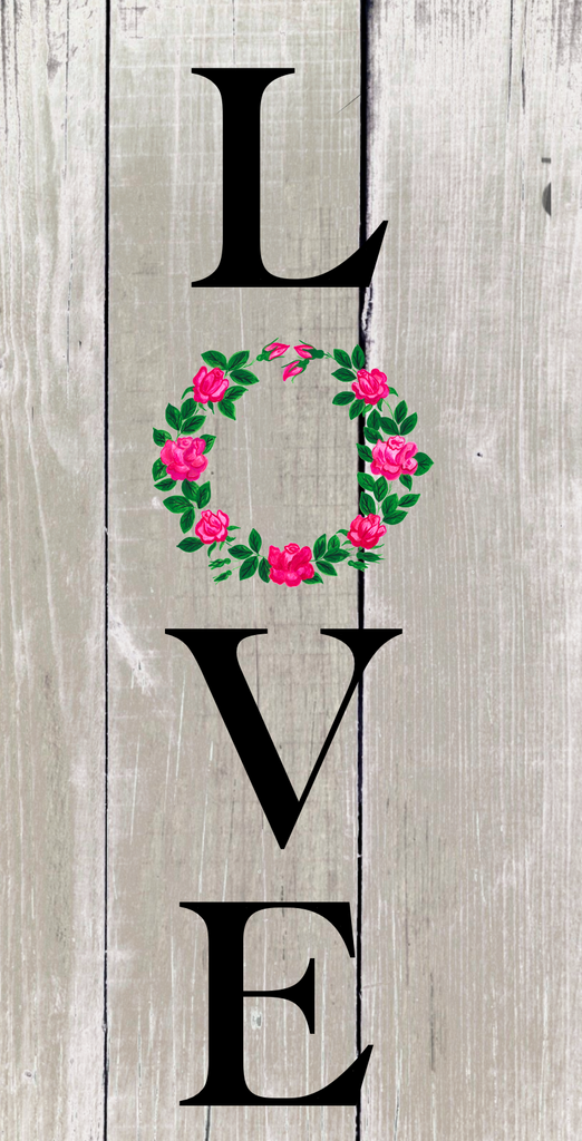 12" metal wreath sign is vertical with the word Love in black letters with the "O" substituted with a simple pink wreath set against a neutral aged wood background.